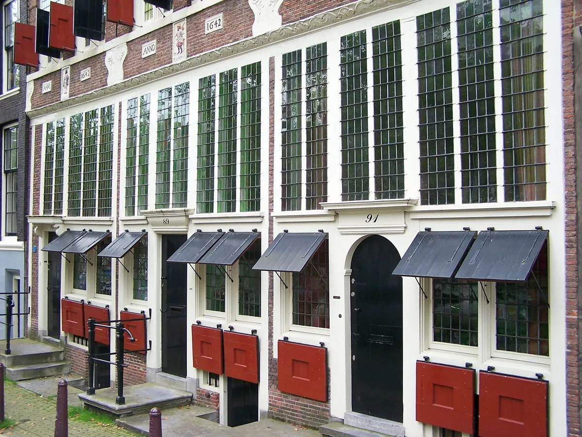 The lower fronts of three white-framed 17th-century houses with leaded windows, black awnings and red shutters, with a carved gable stone and date stones reading ANNO 1642 above