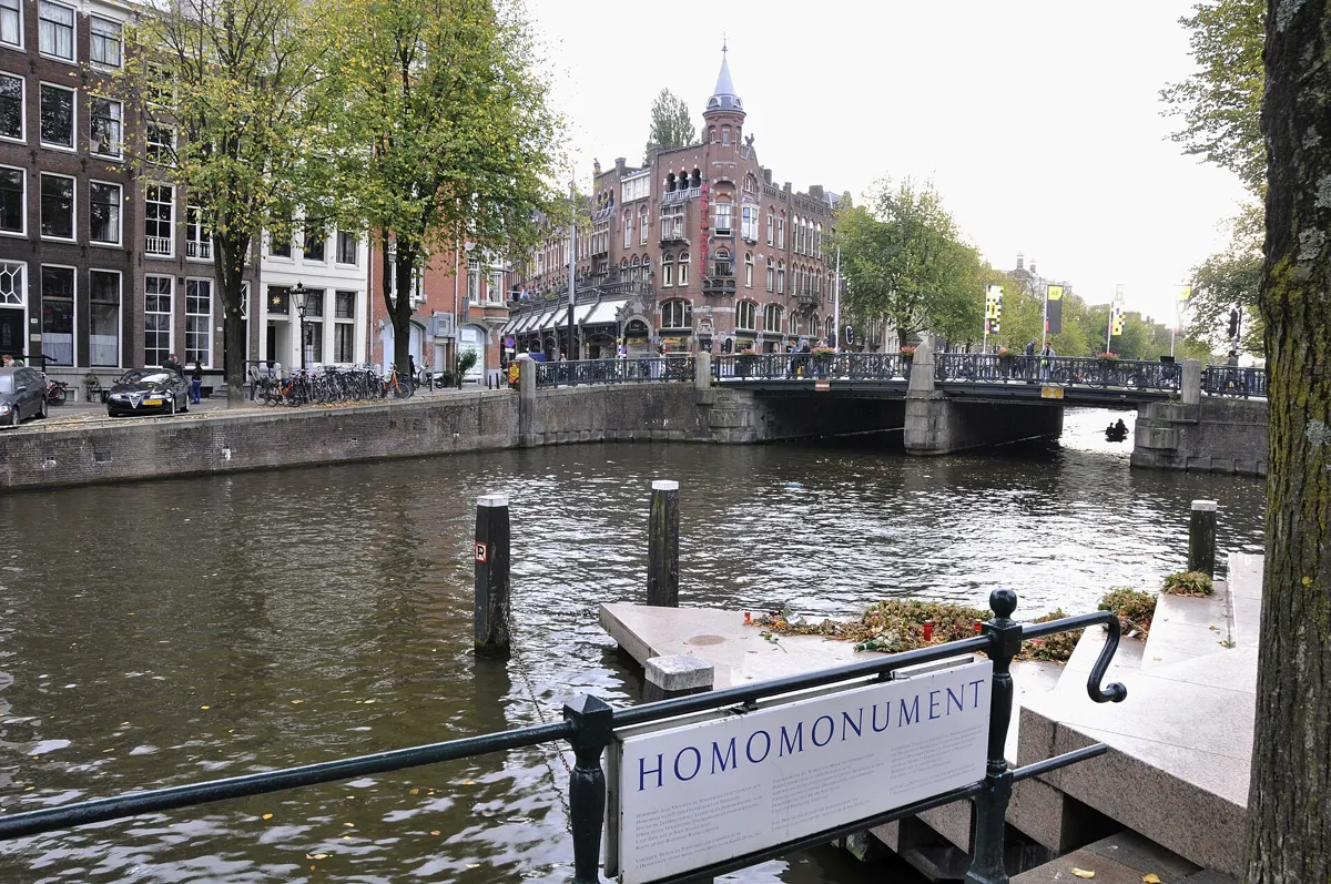 Pink granite steps leading down into the Keizersgracht, with flowers on the platform by the water, a sign reading Homomonument on the railing and a bridge behind