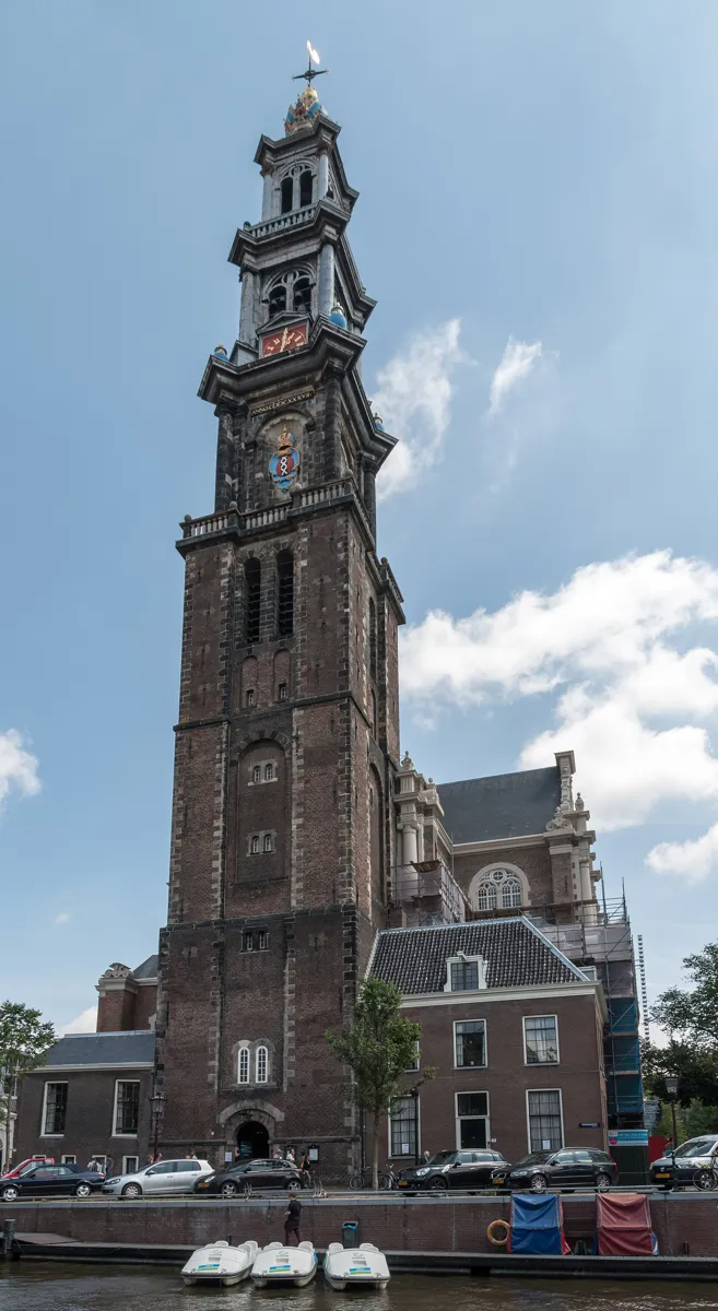 The tall brick and stone tower of the Westerkerk, with a clock, the Amsterdam coat of arms and a crown on top, seen across the Prinsengracht