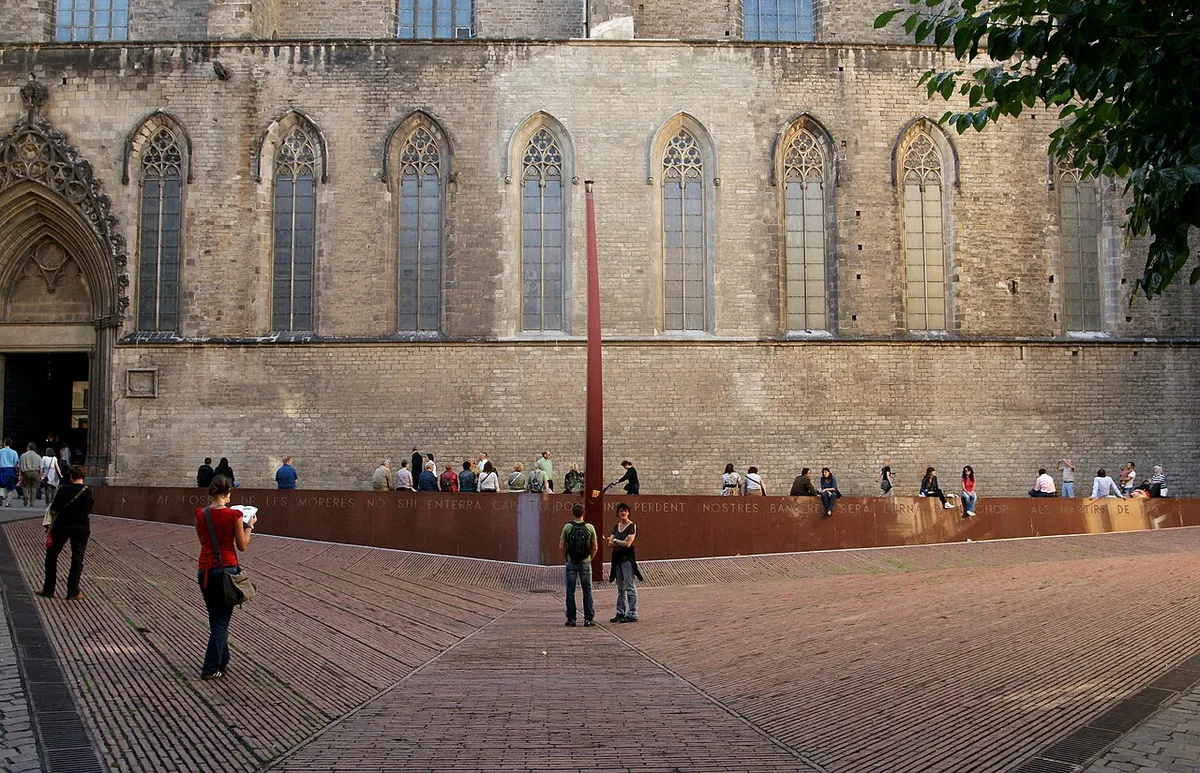The red-brick square of the Fossar de les Moreres, with a long red granite bench and a tall curved column in front of the side wall of Santa Maria del Mar