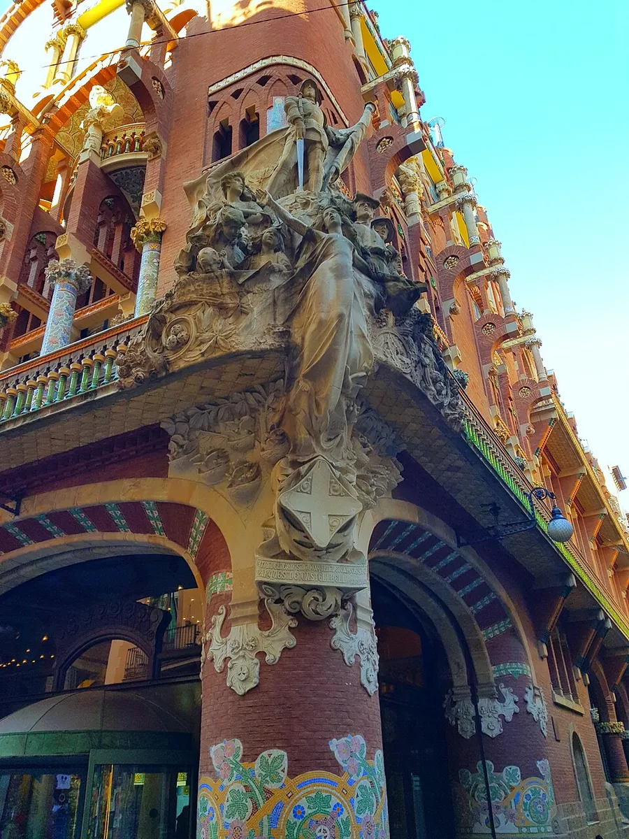 The stone sculpture group on the corner of the Palau de la Música Catalana, a female figure above a crowd of people, over brick columns covered in coloured mosaic