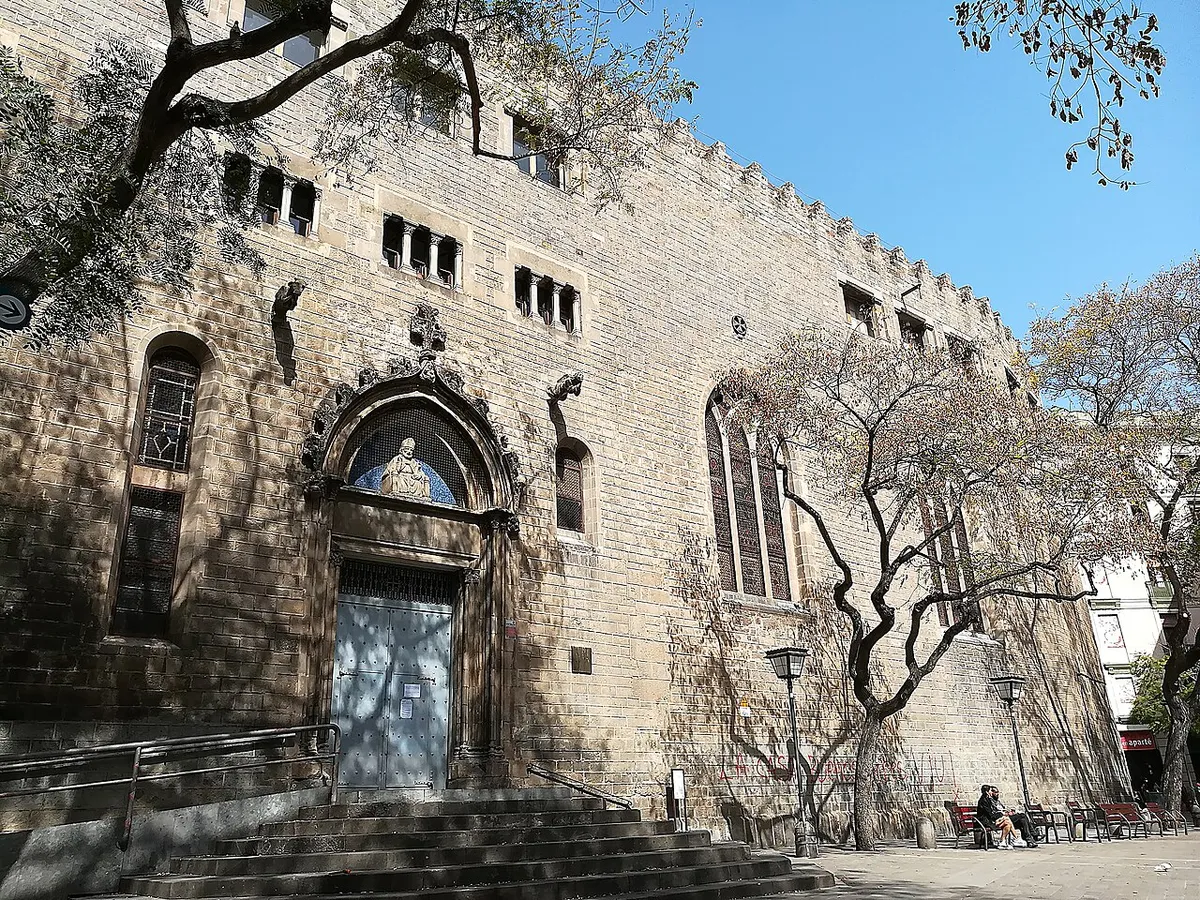 The plain stone front of the church of Sant Pere de les Puel·les, with a Gothic doorway, tall windows and battlements, beside trees in Plaça de Sant Pere