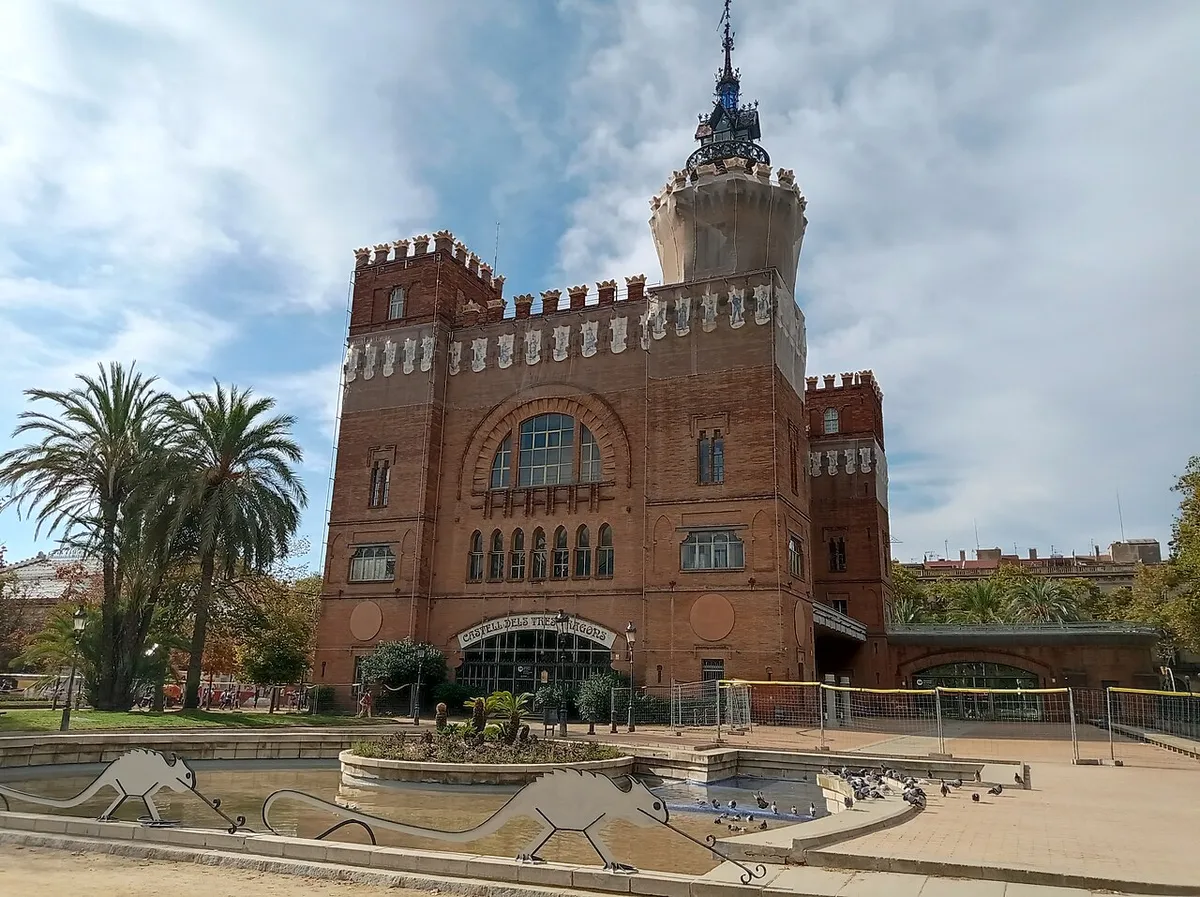 The red-brick Castell dels Tres Dragons in the Parc de la Ciutadella, with battlements, corner towers and a tall spired tower, behind palm trees