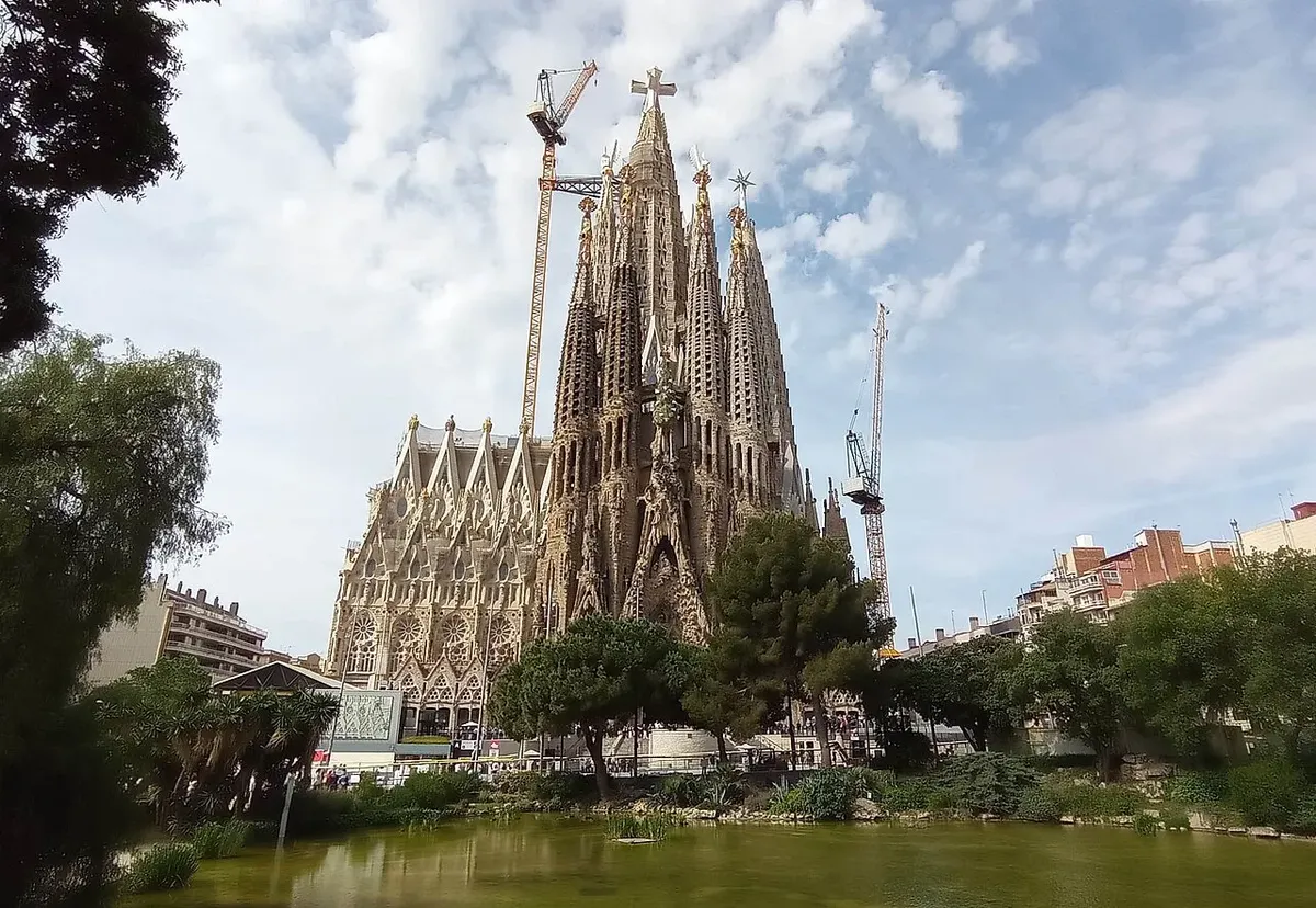 The towers of the Sagrada Família with two construction cranes, seen across a pond and trees