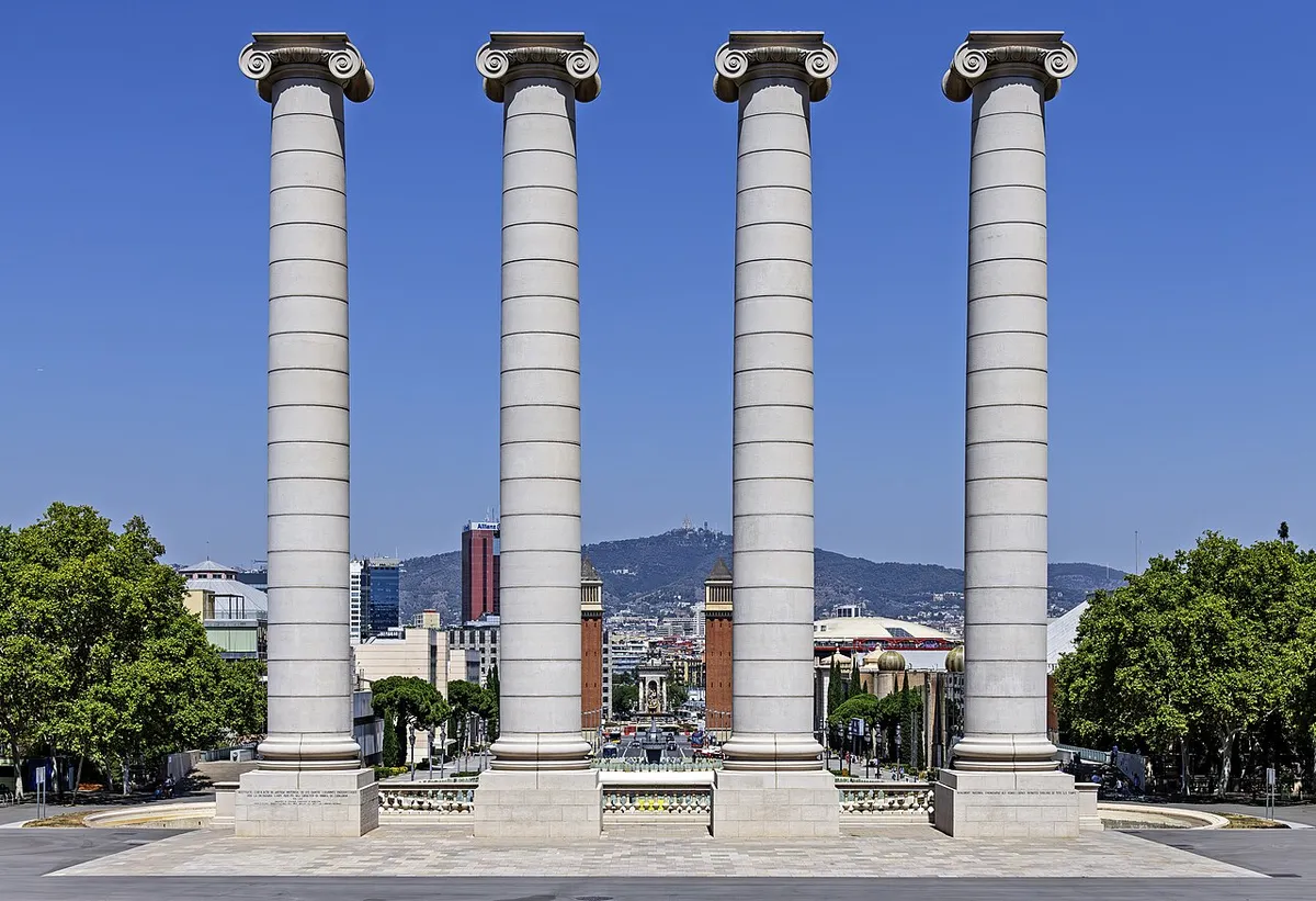 Four tall white Ionic columns on a paved square, looking down towards Plaça d'Espanya and its two brick towers