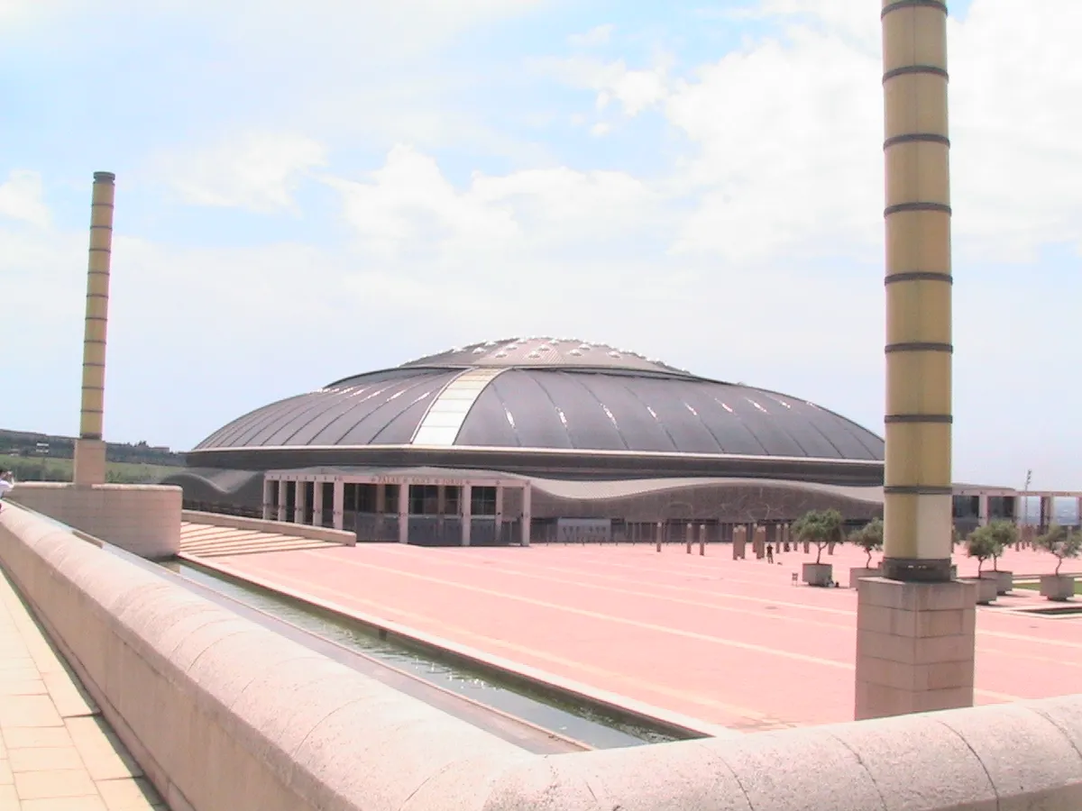 The low dark dome of the Palau Sant Jordi across a wide pink esplanade lined with tall columns