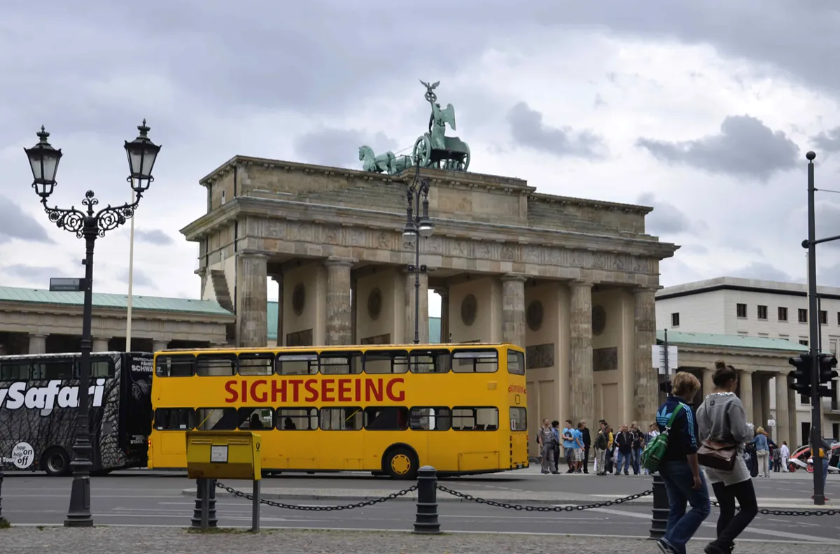 The west side of the Brandenburg Gate, a sandstone gateway of tall columns topped by a chariot with four horses, with a yellow sightseeing bus and people in front