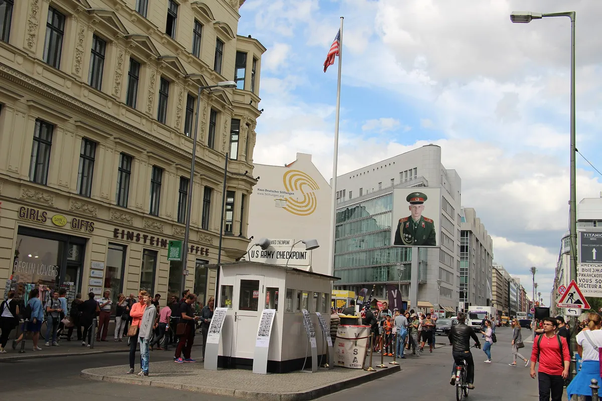 A small white guard hut with an American flag and a sign in the middle of Friedrichstraße, with crowds of tourists, shops and a large backlit portrait of a soldier on a pole behind