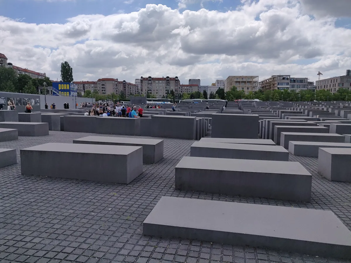 Rows of grey concrete blocks of different heights stretching away across a sloping field, with visitors walking among them and city buildings behind