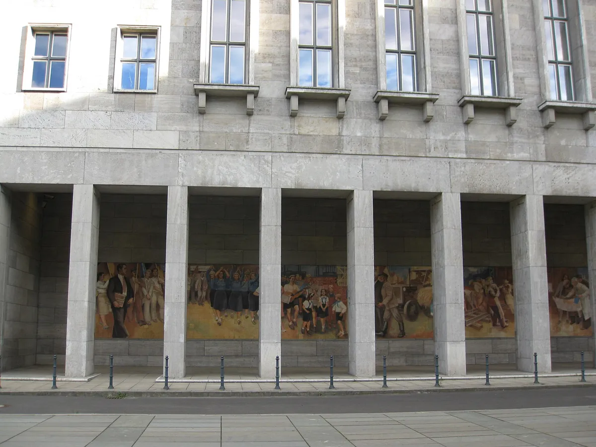 A long colourful painted-tile mural of workers, families and flags, seen through the square stone pillars of a colonnade at the base of a large grey office building