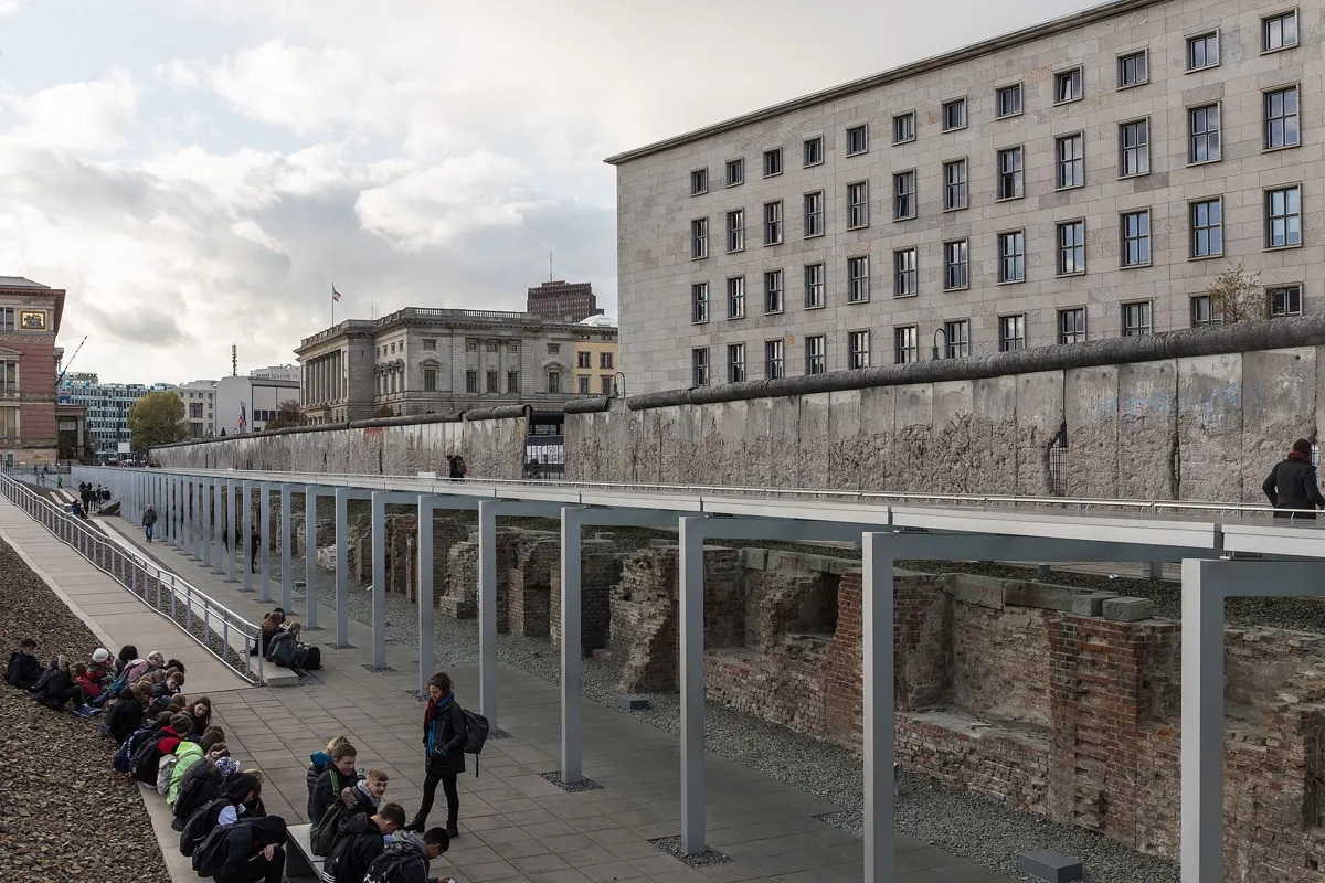A long grey stretch of the Berlin Wall, its surface chipped and full of holes, running above a sunken exhibition trench with old cellar walls, with a large stone office building across the street