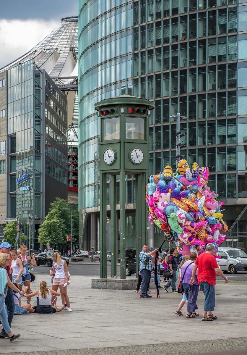 A tall green five-sided tower on steel legs with clocks and traffic lights near the top, standing on a busy pavement in front of glass office towers