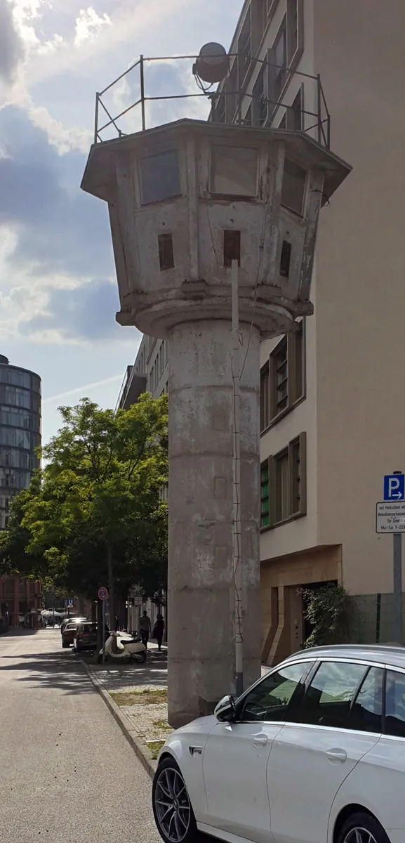 A slim, weathered concrete watchtower with an eight-sided cabin and railing on top, standing on the pavement of a narrow street beside a modern building