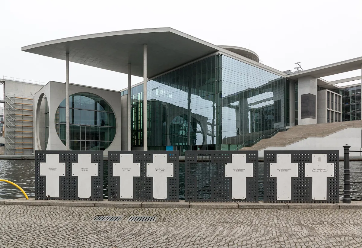 A row of seven white crosses with names on them, fixed to a dark metal railing on the cobbled bank of the Spree, in front of a modern glass and concrete parliament building