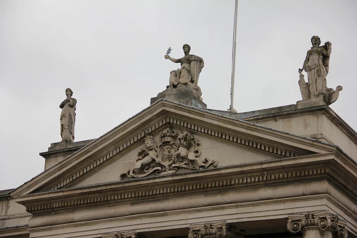 The stone pediment of the Bank of Ireland on College Green, with a carved royal coat of arms and three statues standing on top against a grey sky
