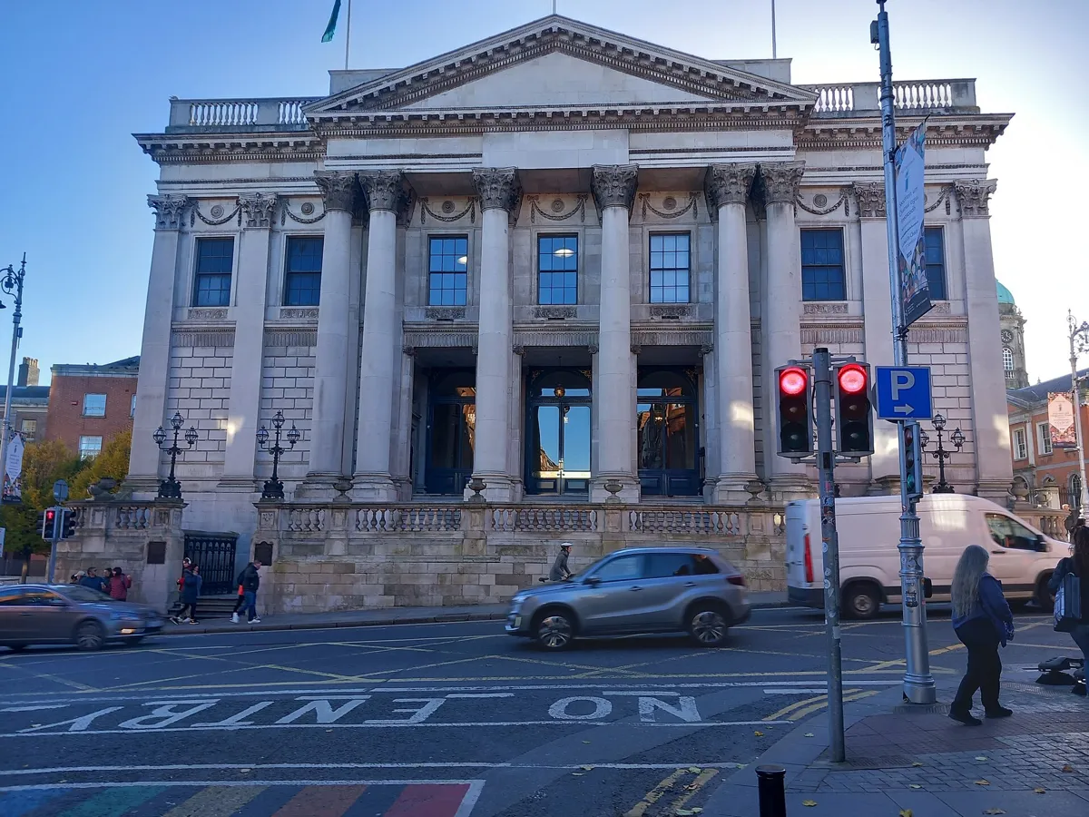 The pale stone front of Dublin City Hall on Dame Street, with six tall columns, a pediment and a balustrade, seen across the road