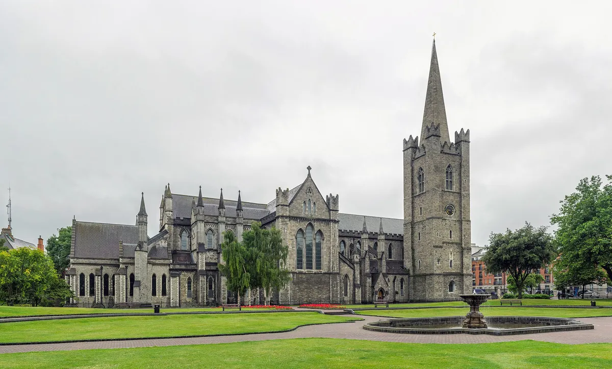 St Patrick's Cathedral, a long grey Gothic church with a square tower and tall spire, seen across the lawns and fountain of St Patrick's Park