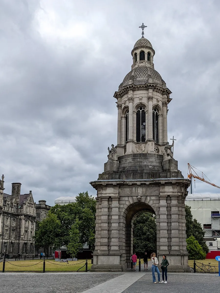 The Campanile of Trinity College, a stone bell tower on an arched base with seated statues at its corners and a small dome on top, standing in a cobbled square