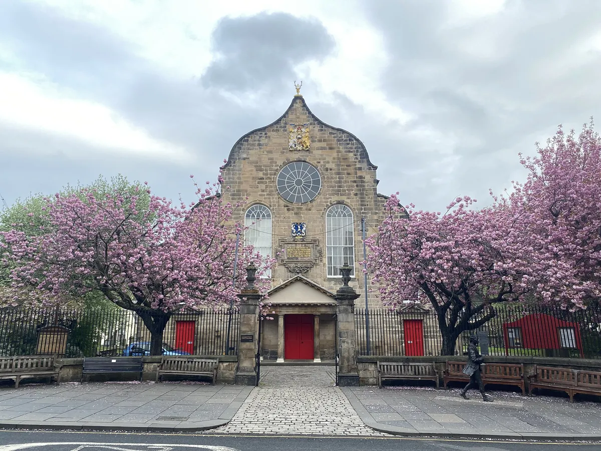 Canongate Kirk, a stone church with a curved Dutch-style gable, a round window, a small columned porch and red doors, framed by pink cherry blossom, with a bronze statue of a walking man on the pavement