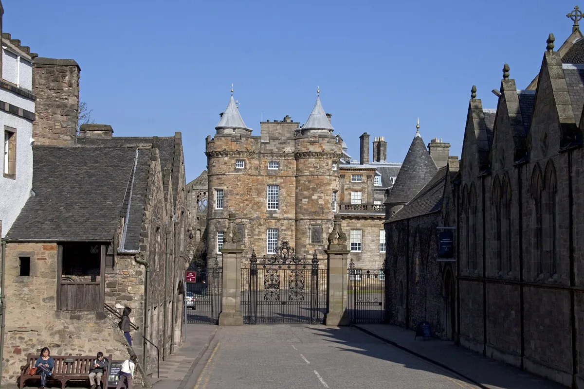 Looking down Abbey Strand to the iron gates of Holyrood Palace, with the palace's old tower and its two pointed turrets behind and old stone buildings on both sides