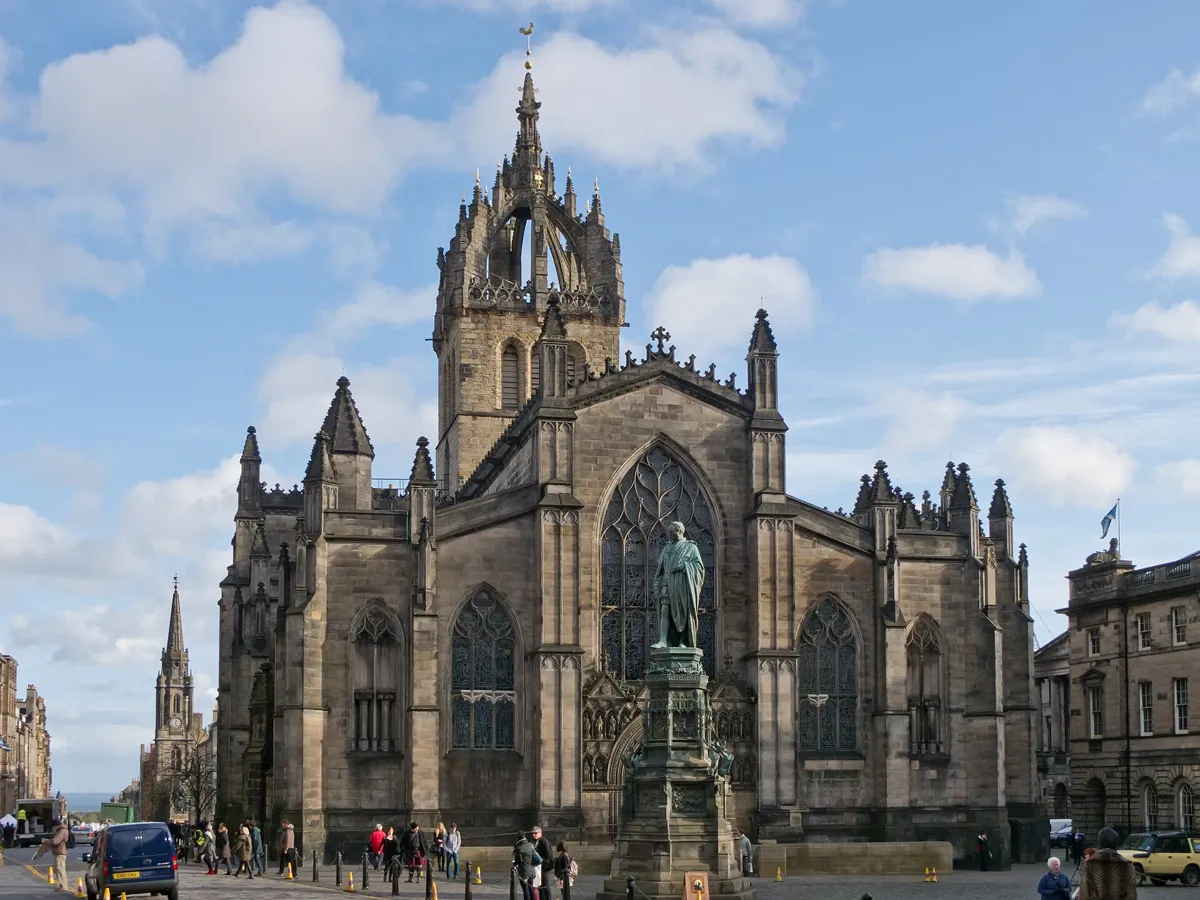 The dark stone west front of St Giles' Cathedral with a large Gothic window and the open crown steeple above, a statue on a tall pedestal in front and people on the cobbles