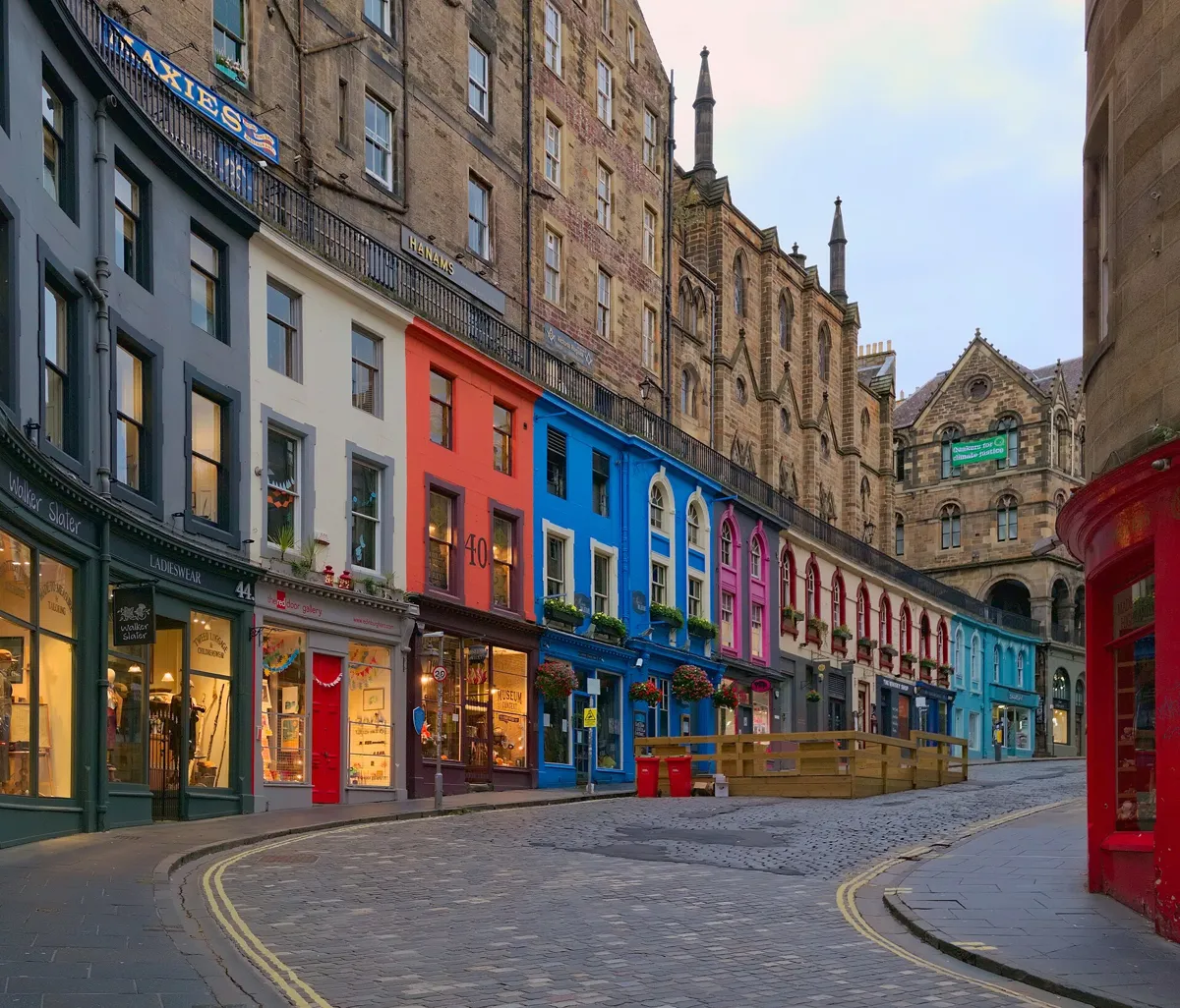 The curve of cobbled Victoria Street, lined with shopfronts painted grey, white, red, blue and pink, with taller stone buildings and a railed terrace above them