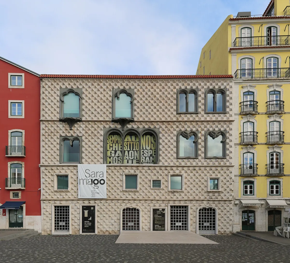 The front of the Casa dos Bicos, a four-storey house covered in rows of pointed pale stones, with arched upper windows, between a red and a yellow building