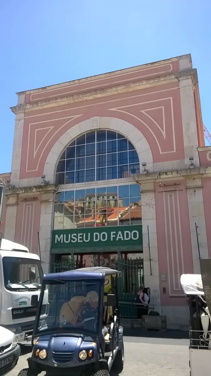 The pink front of the Museu do Fado, with a tall arched window, a green sign reading MUSEU DO FADO over the door and a small electric tourist cart parked outside