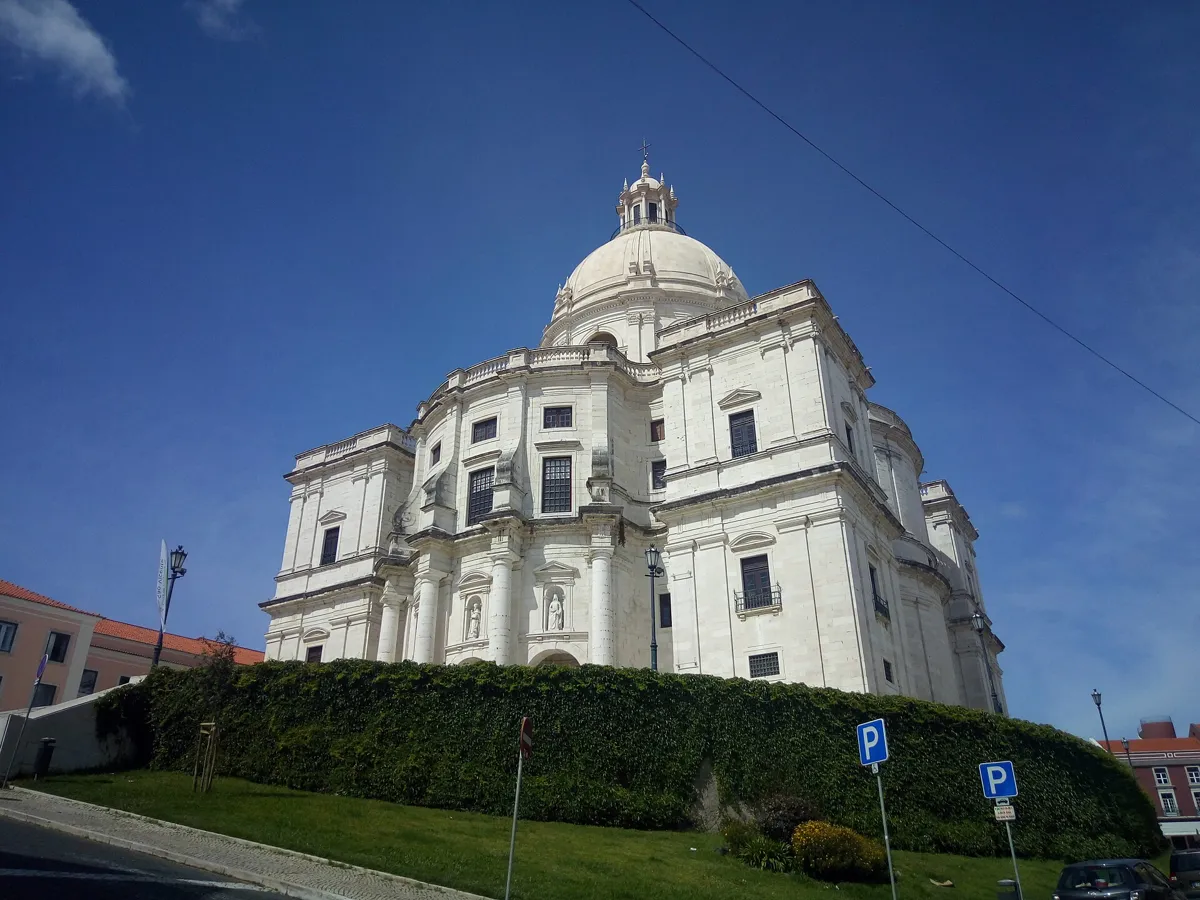 The white National Pantheon, a church with curved walls, corner towers and a large white dome, seen from below a green hedge under a blue sky