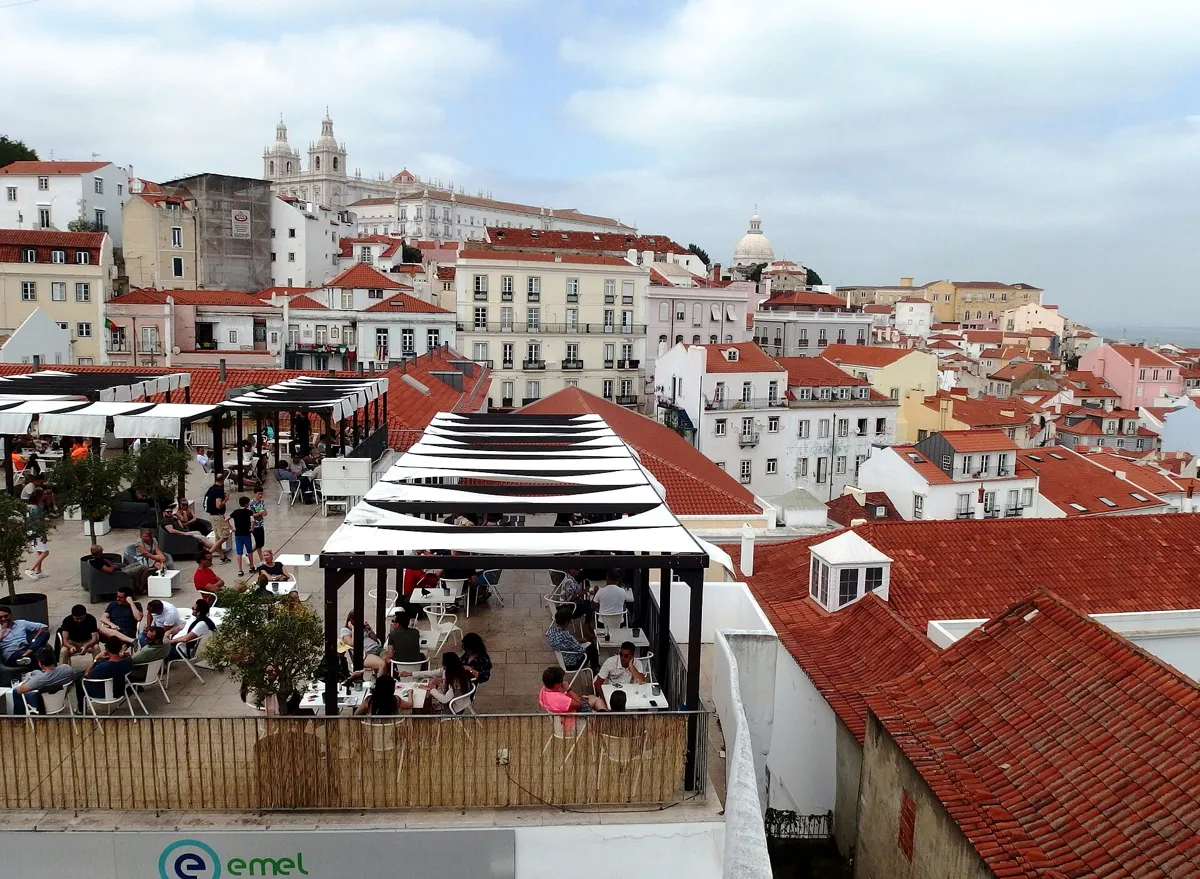 View from the Portas do Sol terrace over a café with white awnings and red-tiled Alfama roofs, with the white towers of São Vicente de Fora on the hill behind