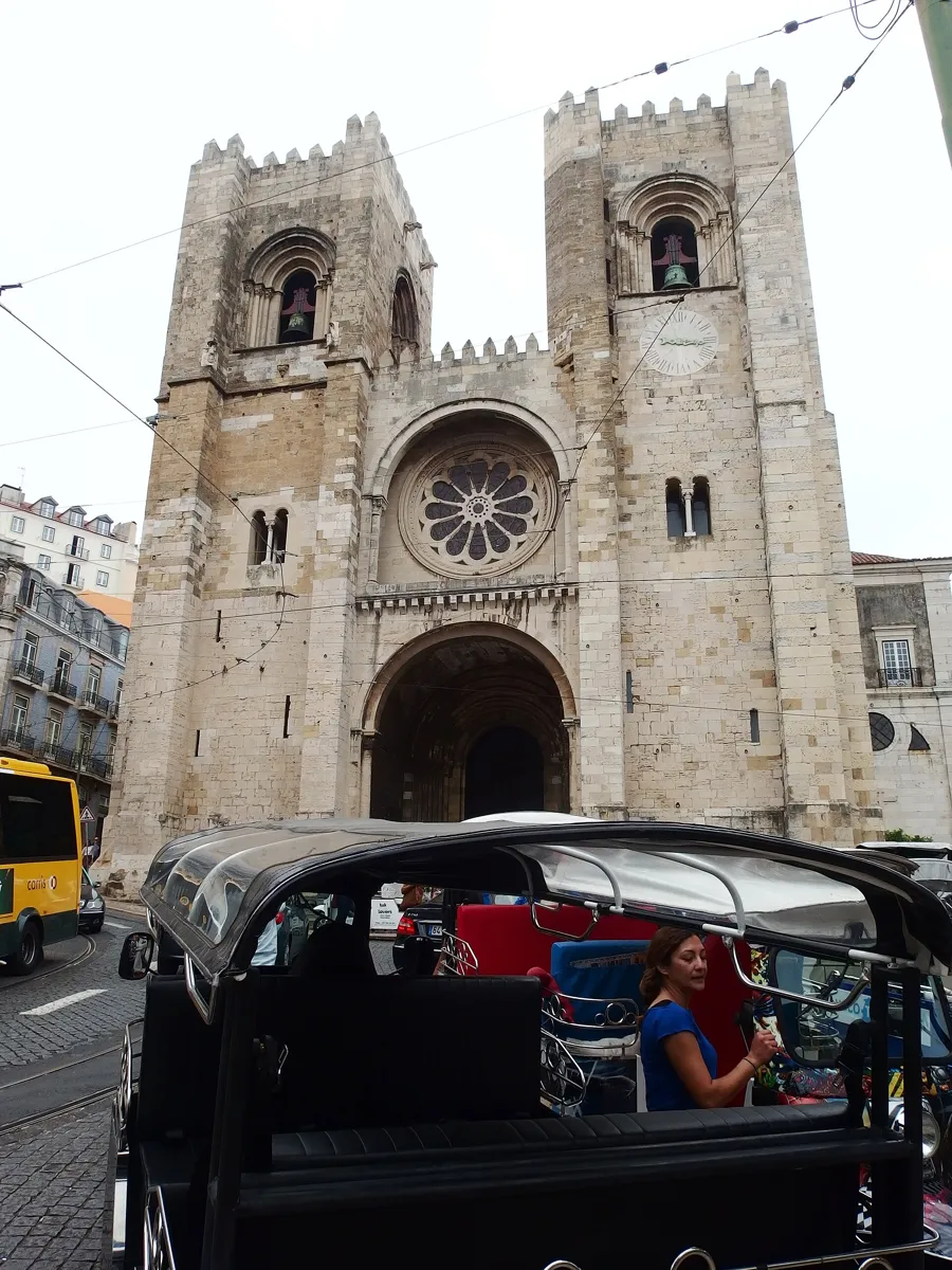 The west front of Lisbon Cathedral: two square stone towers with battlements, a round rose window between them and a deep arched doorway, with a tuk-tuk parked in front