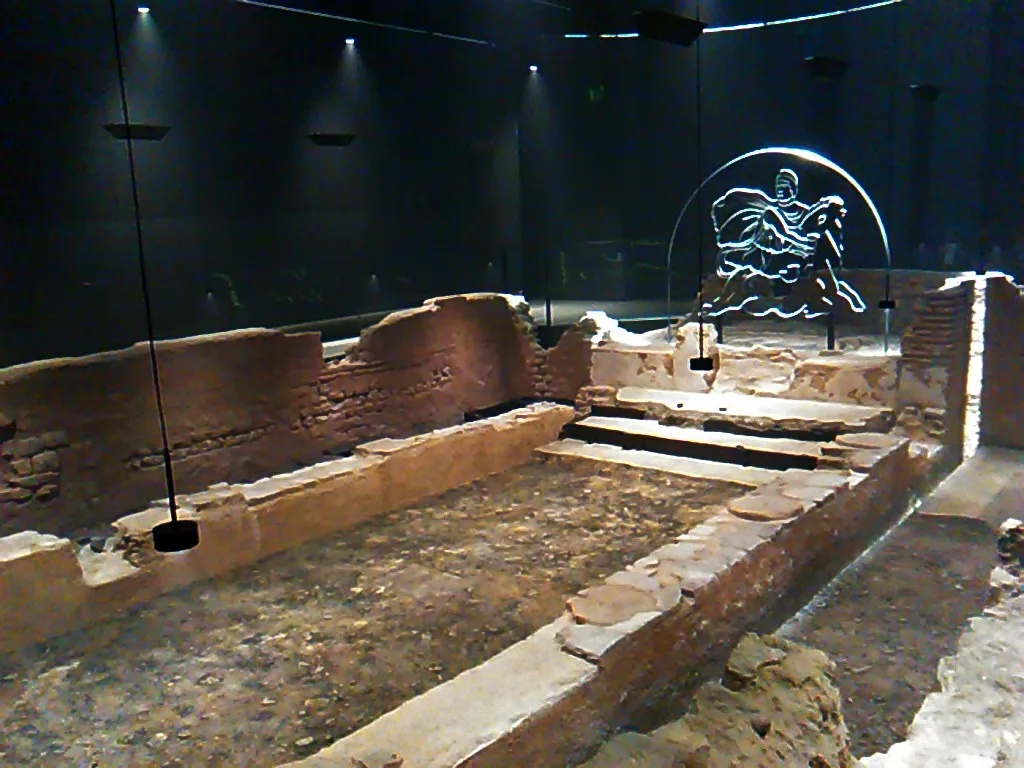 The low stone and tile walls of the Roman temple of Mithras in a dark underground room, lit by spotlights, with a glowing outline of Mithras killing a bull at the far end