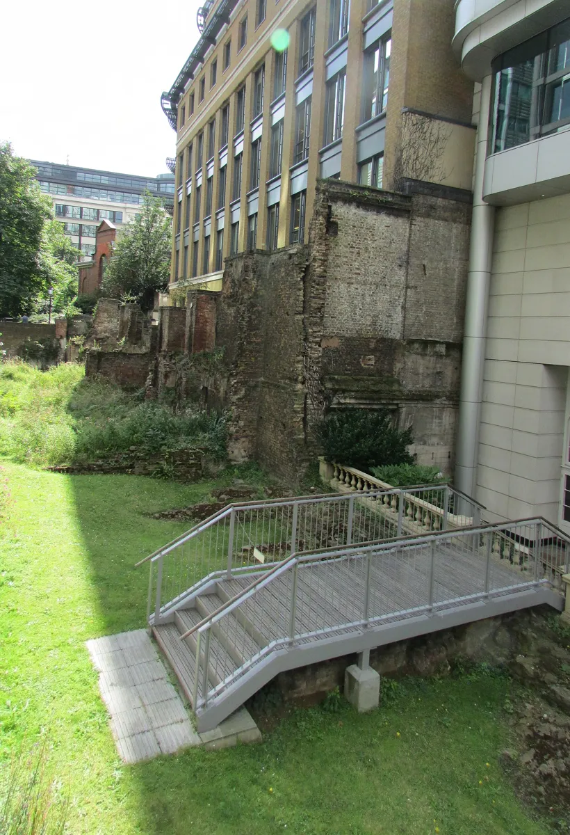A long ruined wall of stone and old brick in a sunken lawn beside modern office buildings, with a metal footbridge and steps in the foreground