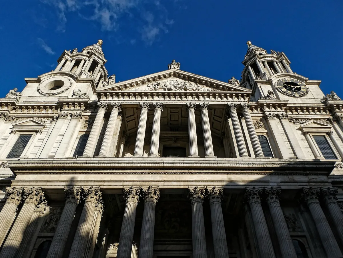 The west front of St Paul's Cathedral seen from below: two tiers of paired stone columns, a carved pediment and two baroque towers, one with a clock, against a blue sky