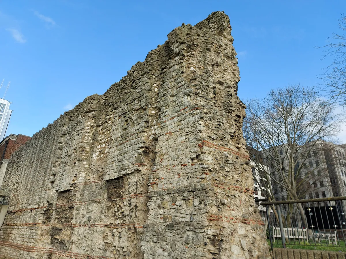 A tall ruined wall of rough grey ragstone with thin horizontal bands of red tile in its lower part, seen from below against a blue sky, with railings and bare trees