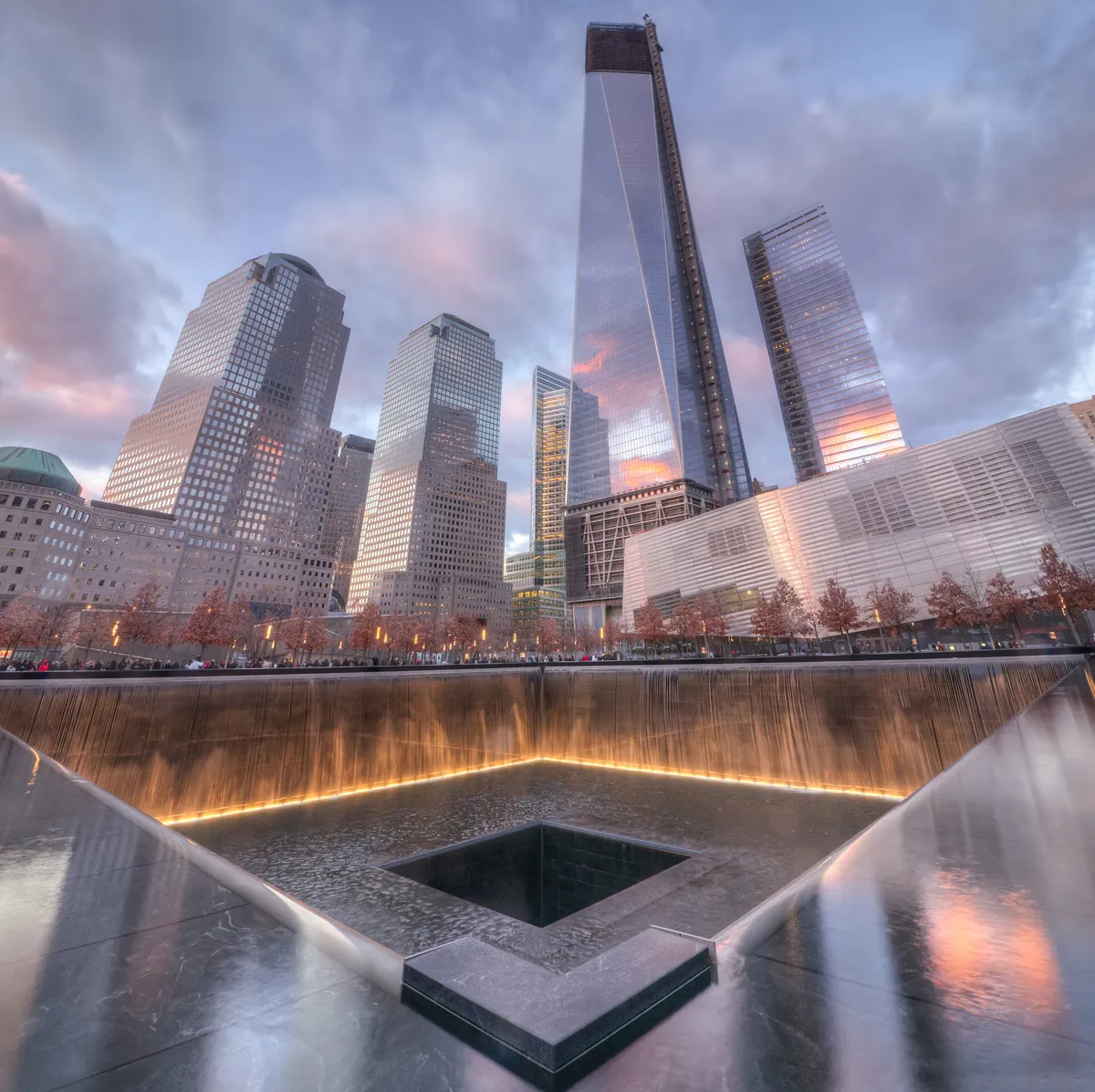 The corner of a 9/11 Memorial pool at dusk, with water falling down lit black walls into a square basin and a smaller dark void, and tall glass towers behind