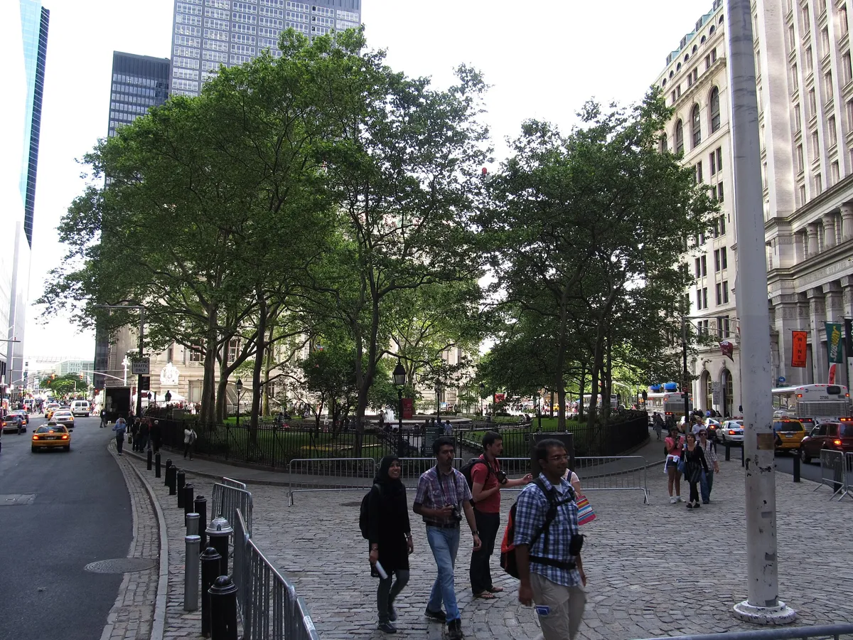 Bowling Green seen from the cobbled plaza to the north, a small park of tall trees inside a low black iron fence, with pedestrians in front and stone office buildings around it