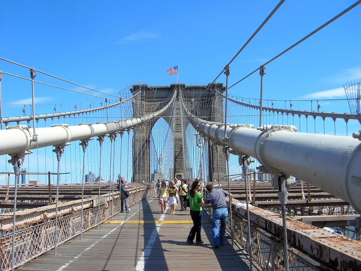 People walking on the wooden promenade of the Brooklyn Bridge between thick suspension cables, towards a stone tower with two pointed arches and an American flag