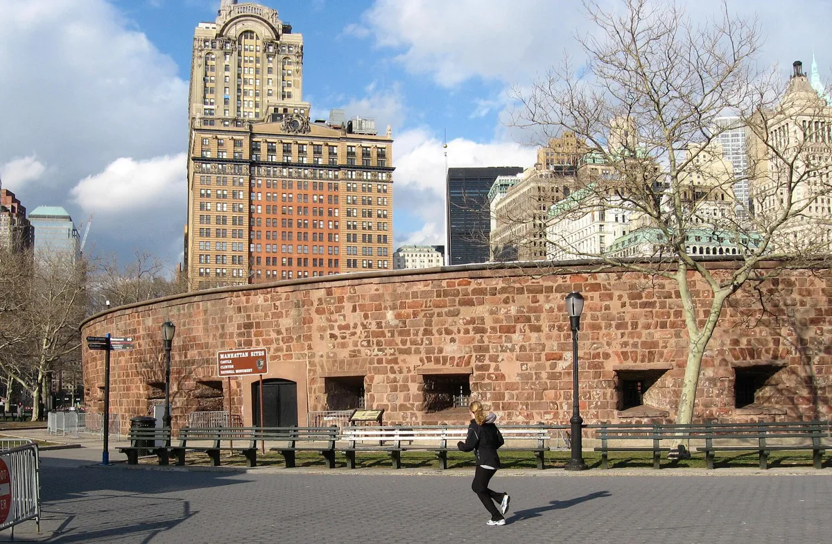 The low round wall of Castle Clinton in red sandstone, with a doorway, small gun openings and benches in front, and tall office buildings behind