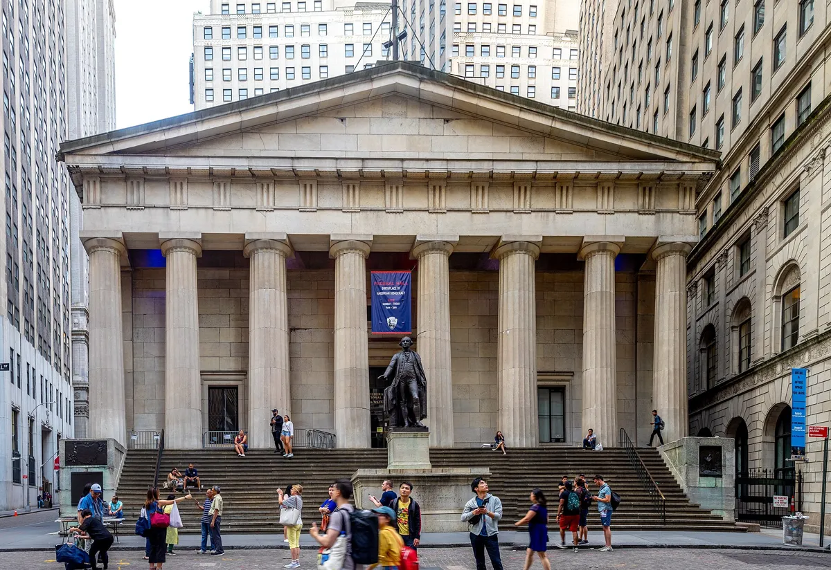 Federal Hall, a marble building like a Greek temple with eight Doric columns and a wide flight of steps, with a bronze statue of George Washington in front and visitors on the steps