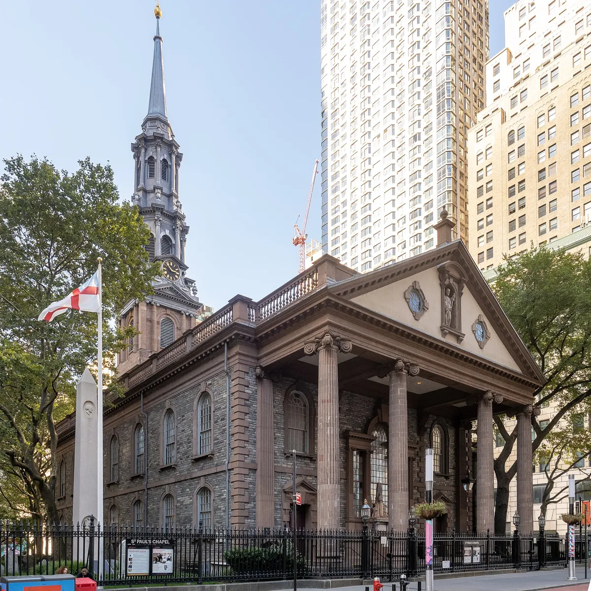 St. Paul's Chapel from Broadway, a stone church with a columned portico, a statue in a niche in the pediment and a tall spire, behind an iron railing, with an obelisk and a flag in the churchyard