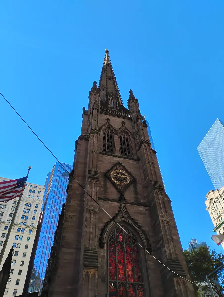 The dark brownstone Gothic tower and spire of Trinity Church seen from below, with a clock, a tall red stained-glass window and glass office towers behind