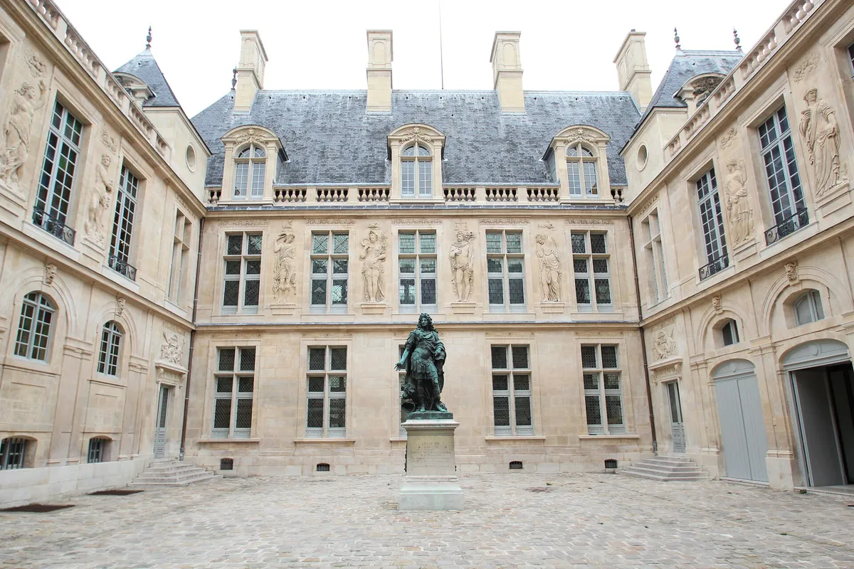 A paved courtyard enclosed by pale stone wings carved with tall figures, with a dark statue of Louis XIV in Roman dress on a plinth in the middle