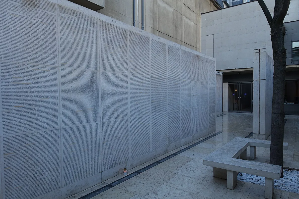 Pale stone walls engraved with thousands of names in small letters, with a stone bench and a young tree in the courtyard