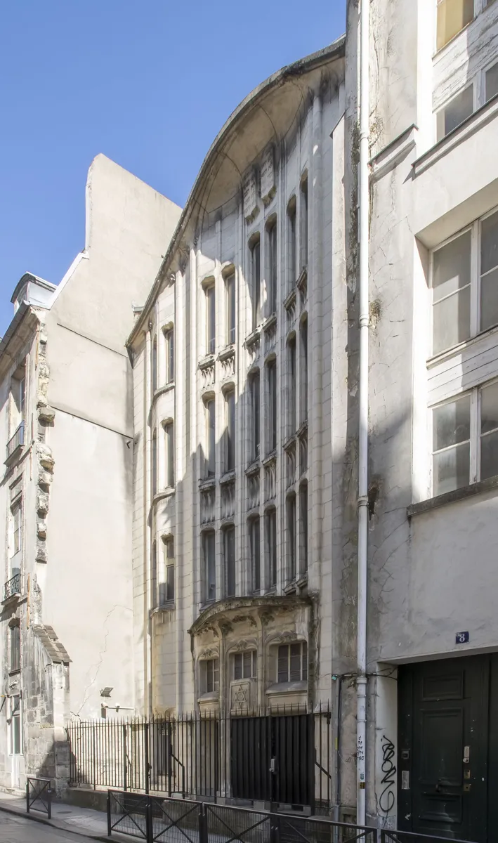 A tall, narrow pale synagogue front with thin vertical piers, slit windows and a wavy curved top, behind iron railings on a narrow street