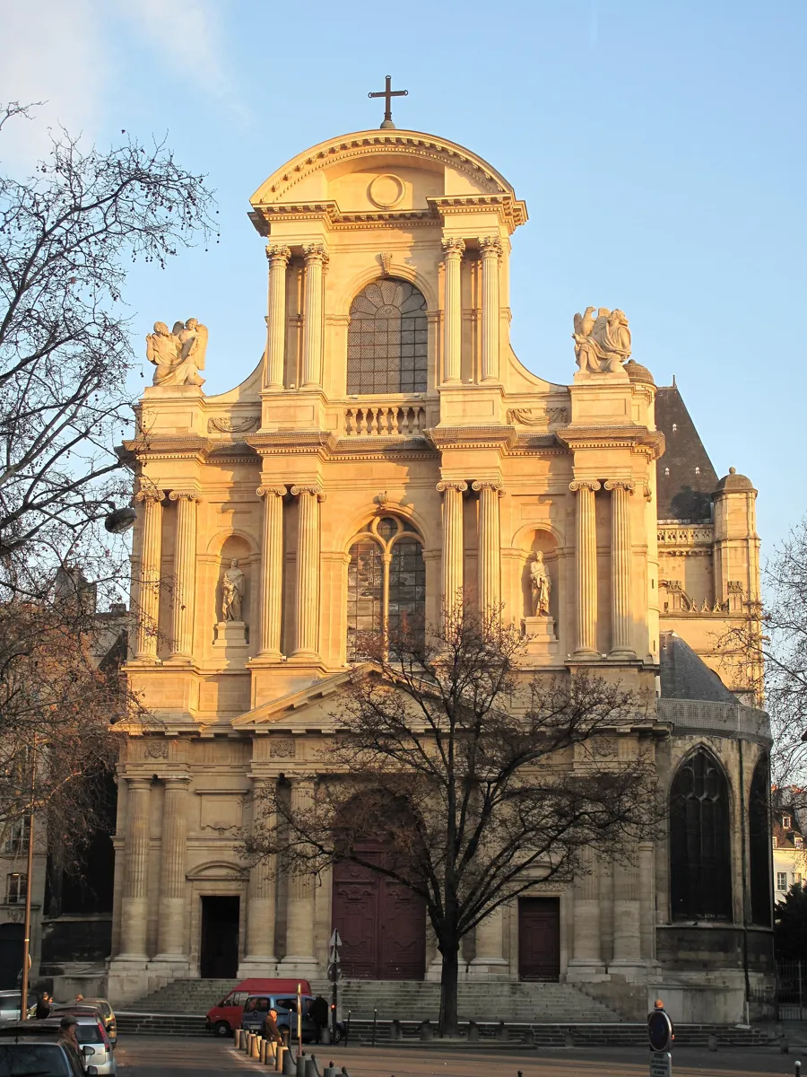 The tall pale stone front of Saint-Gervais, with three tiers of paired columns, statues in niches and a curved pediment, behind a bare tree on the square