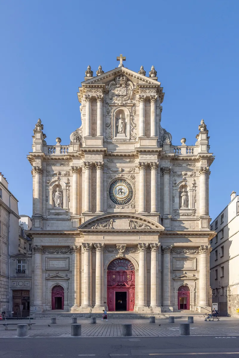 The tall, richly carved stone front of Saint-Paul-Saint-Louis, with stacked columns, statues in niches, a round clock and red doors, under a clear blue sky