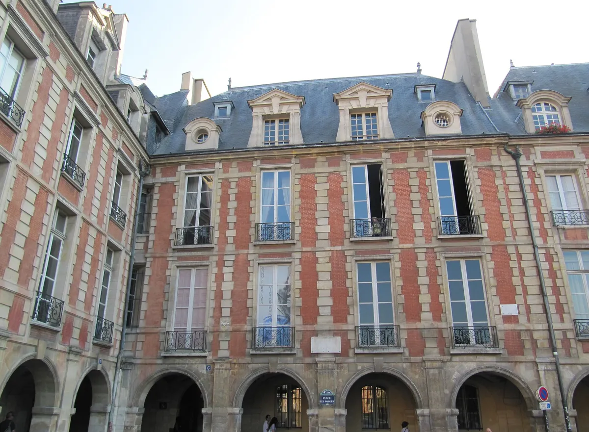 Red brick house fronts with pale stone bands, iron balconies and slate roofs with dormers above stone arcades, with a Place des Vosges street sign under the arches