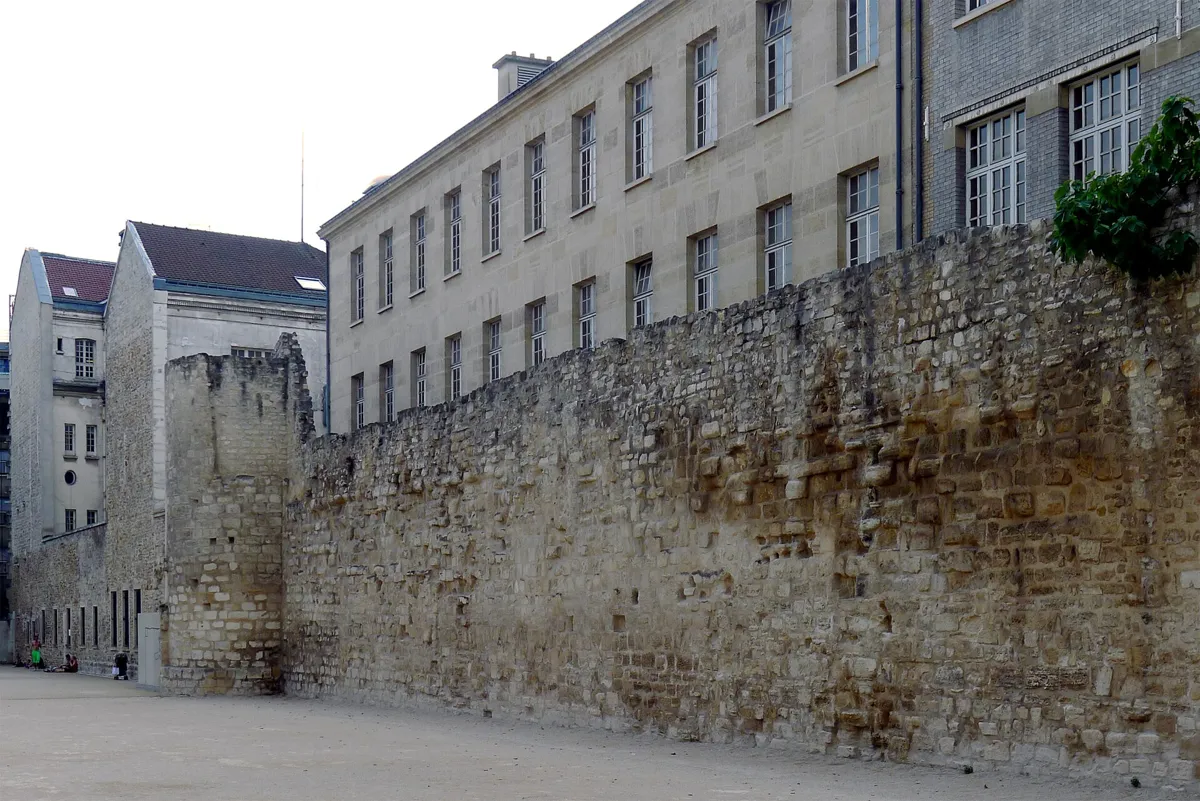 A long, high wall of rough medieval stone with a squat tower, beside a gravel sports ground and the plain windows of school buildings behind