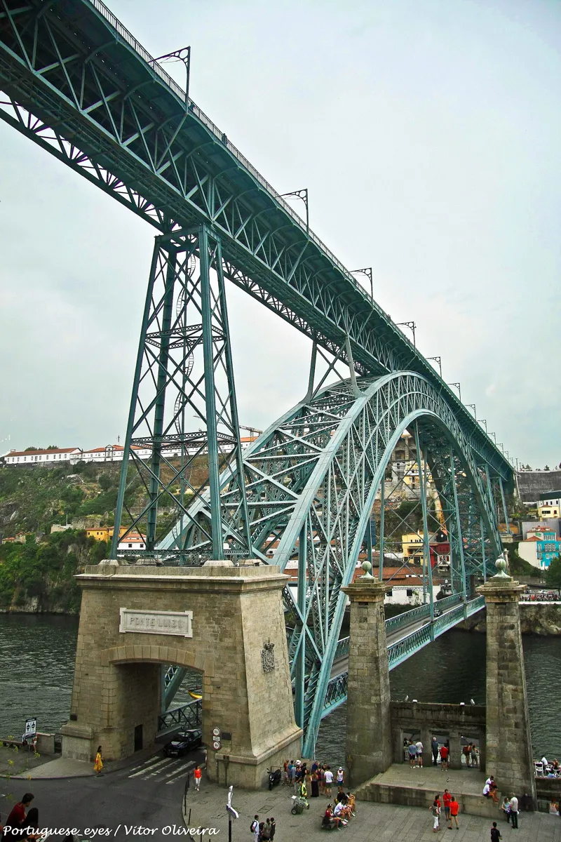 The iron arch of the Luís I bridge seen from the Porto end, with the high upper deck above, the lower deck crossing the river, and a stone gateway marked PONTE LUIZ I